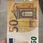 Купюра в 50 євро між пальців на сірому фоні — символ готівкової валюти