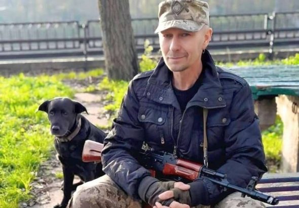 Сергій Русінов сидить на лавці з автоматом у руках, поруч стоїть собака